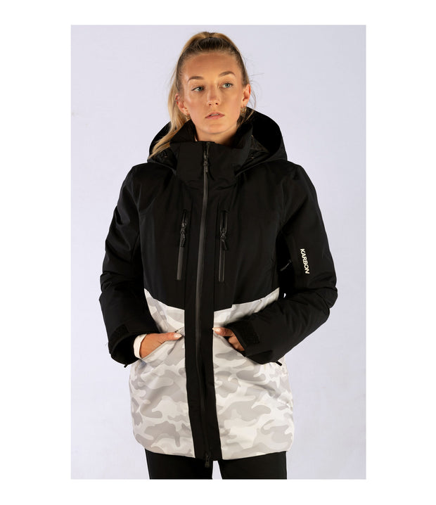 A woman wearing a two-tone insulated jacket with a black hood, shoulders, upper chest, and upper sleeves, paired with a camo pattern body and lower sleeves. The jacket features a front zip closure, two zippered chest pockets, and two side zip pockets, with an adjustable hood.