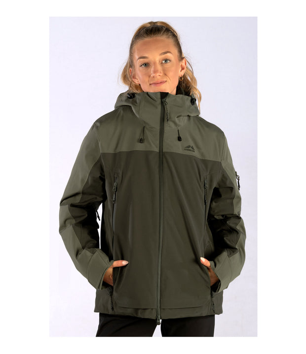 A woman wearing a two-tone shell jacket with a lighter green hood, shoulders, and upper chest, paired with a darker green body and sleeves. The jacket features a front zip closure, zippered chest pocket, and two side zip pockets, with an adjustable hood.
