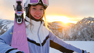 Person holding a pink snowboard with a sunset and snowy landscape in the background

