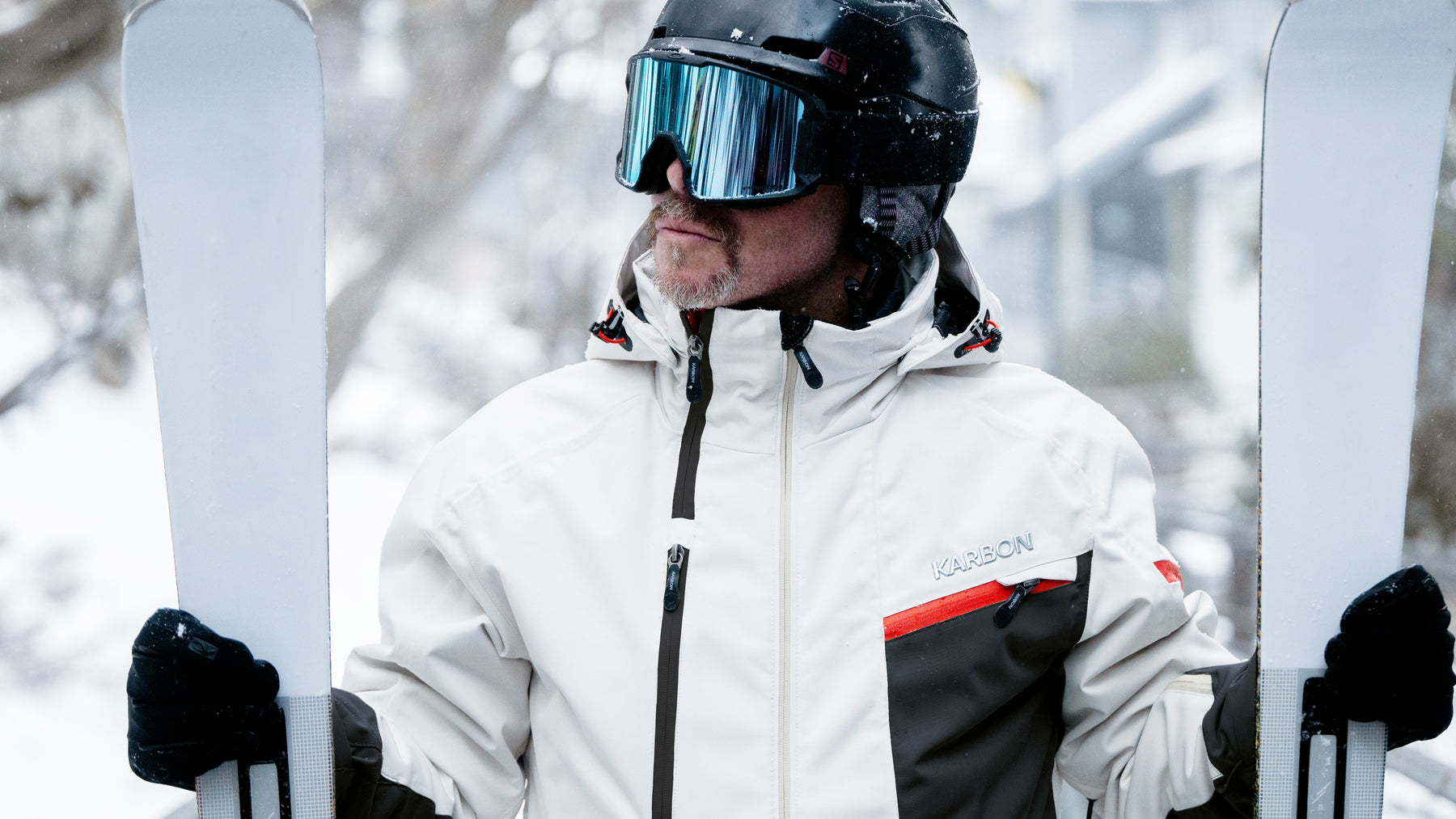 Person in ski gear holding skis with a blurred snowy background


